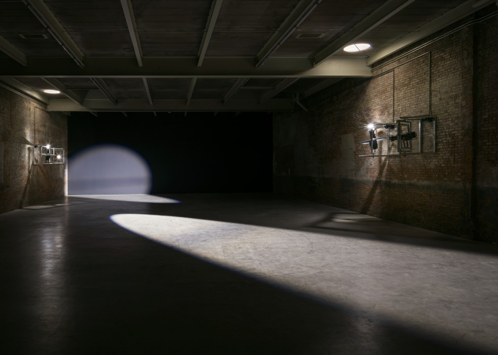 A dark room with cement floors, brick walls on the left and right and a black wall in the background are lit with two spotlights, one seen mounted on the wall on the right, the other mounted on the left wall.